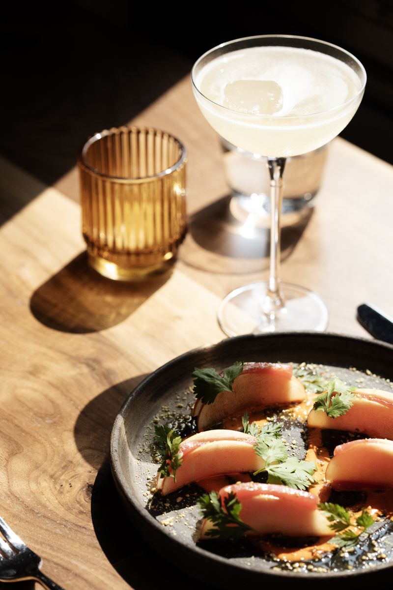 A black plate with small piece of fish garnished with herbs next to a gimlet glass. 