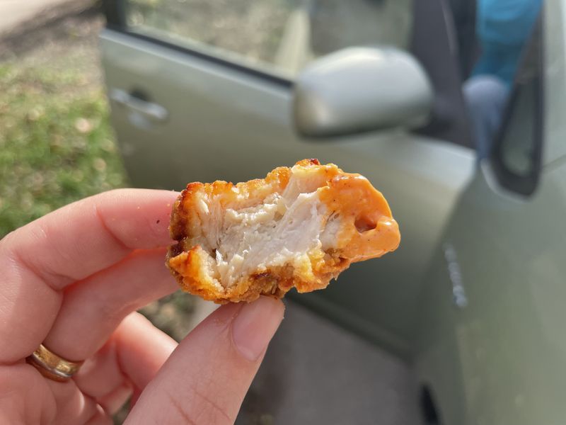 A hand holding a bitten-into chicken nugget with thick breading, white meat inside, and red sauce on the right side in front of an open car door. 