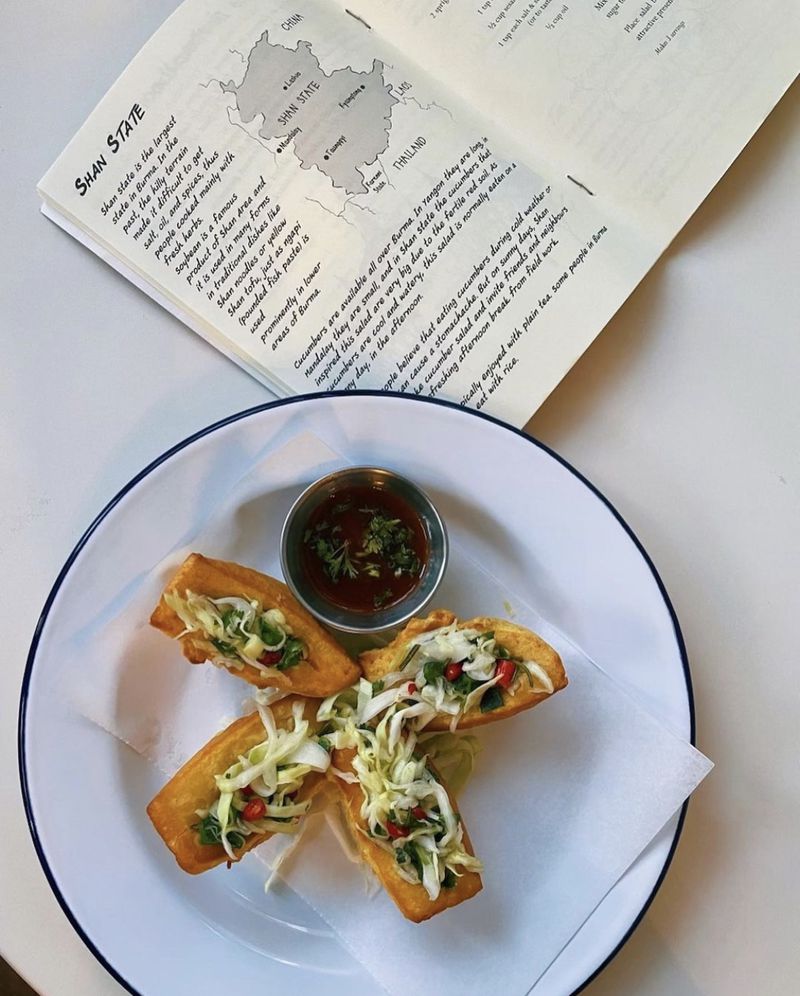 Four pieces of crispy tofu topped with a veggie slaw on a round white plate, on a white table, next to an open book that reads “Shan State” with a drawing of a map. 