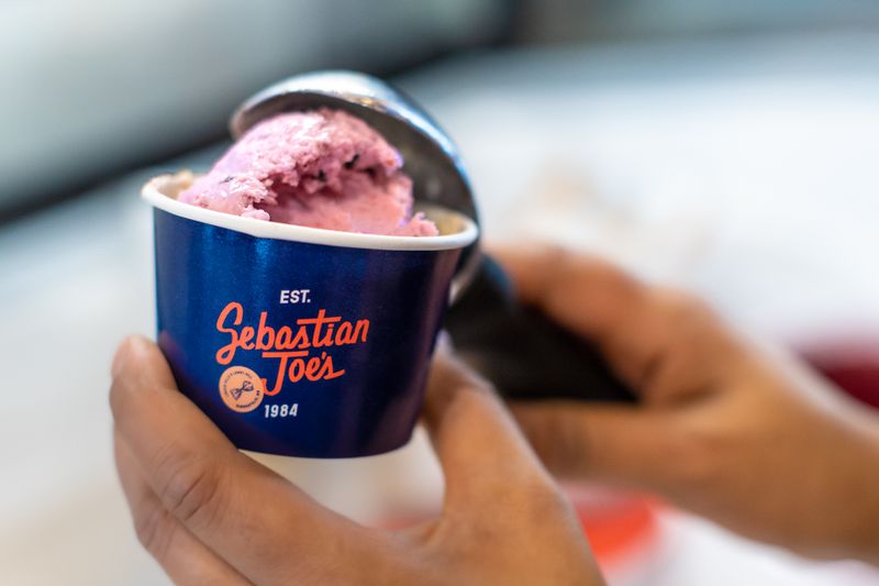 A blue ice cream dish with the words “Sebastian Joes Est. 1984” on it, and a hand scooping pink ice cream into it. 