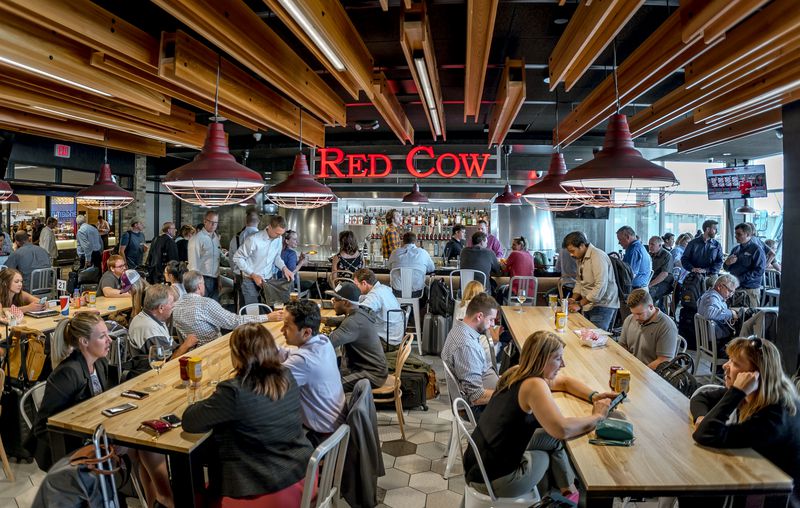 The interior of an airport restaurant, with many people sitting at long wooden tables drinking beer, and a red sign in the background that says “Red Cow.”