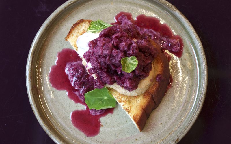 A beige stone plate with a half-slice of milk toast, topped with sesame mousse and purple concord grape ice cream. 