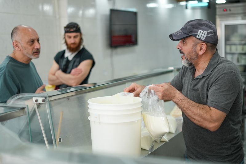 Two men standing on one side of a glass counter talking to a man standing behind the counter filtering yogurt into a white plastic bucket, wearing a grey shirt and a grey hat. 