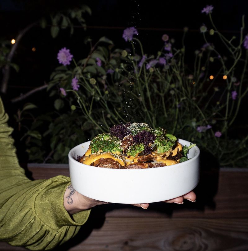A hand in a silky green sleeve holding a white dish full of potato skins and broccoli, drizzled with an orange sauce, with flowers in the background. 