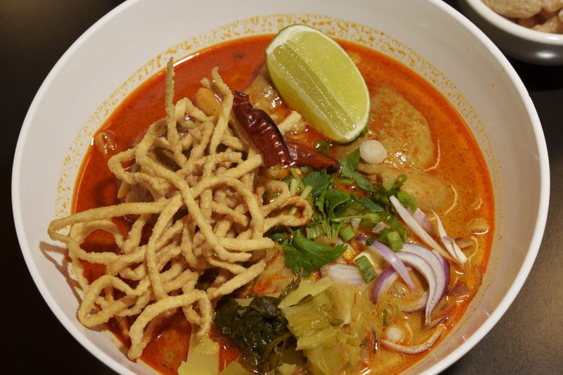 A white bowl of bright red and orange khao soi soup, topped with chilis, onions, a wedge of lime, cilantro, and crispy fried noodles. 