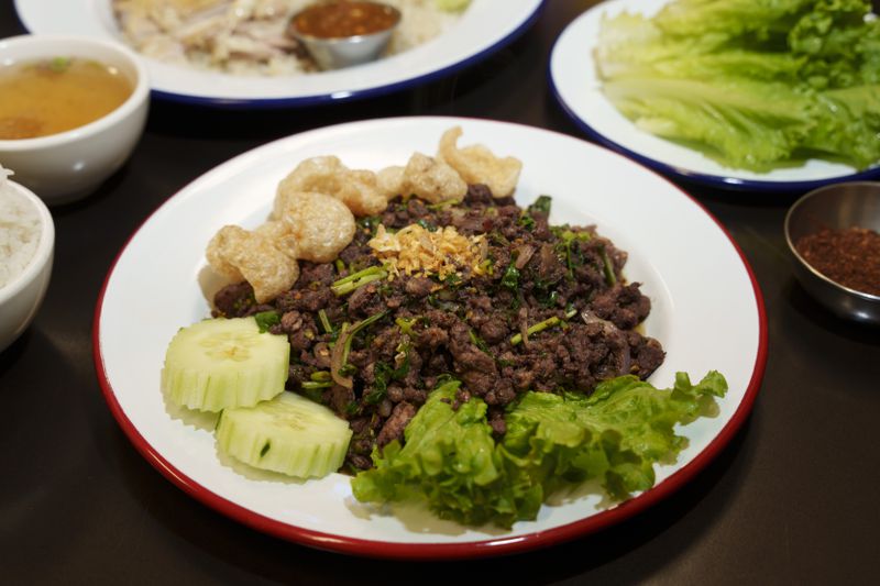 A white, red-rimmed plate of beef larb with pork rinds, two slices of cucumber, and a leaf of lettuce on a black table, with other dishes visible in the background.