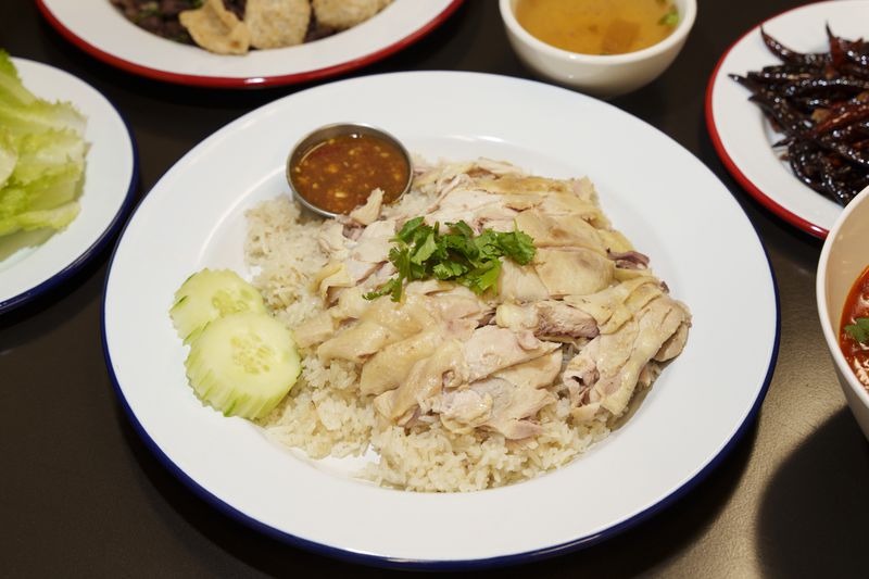 A white plate of chicken, rice, two cucumber slices, and red sauce on a black table, with other dishes visible in the background. 