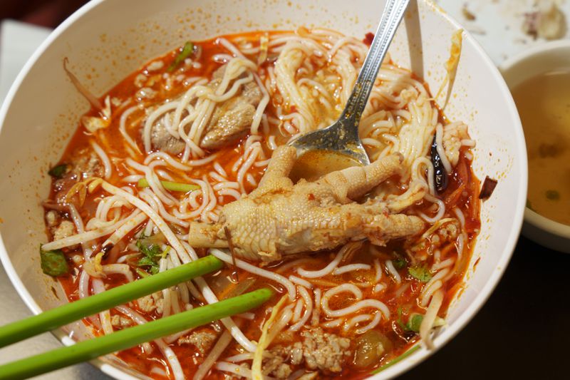 A white bowl of bright red noodle soup with chiles and a chicken foot in it, plus two green chopsticks and a spoon. 