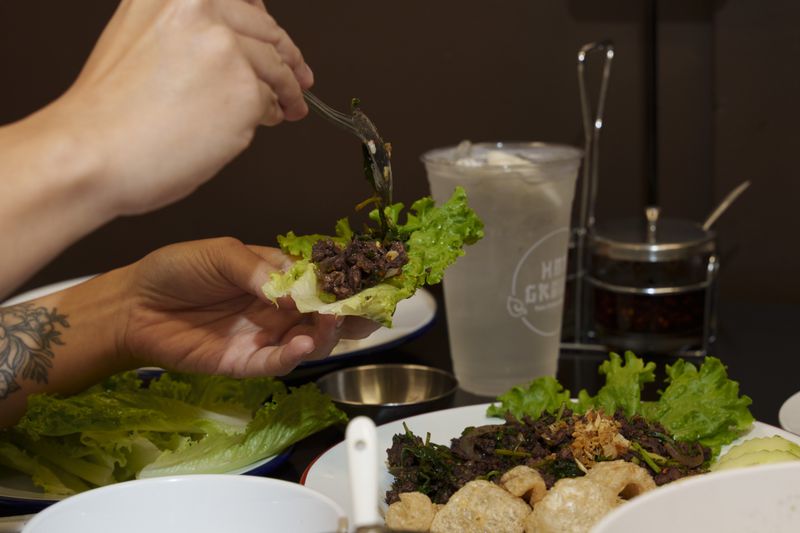 Two hands spooning beef larb onto a piece of lettuce above a table set with food and a drink. 