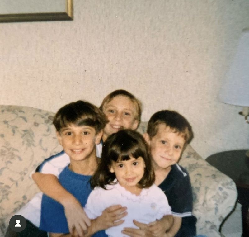 A young girl with brown hair and three boys with brown hair all hugging each other and sitting on a couch in front of a white wall.