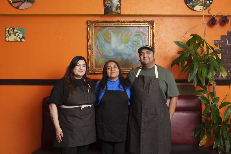 Diana and Miguel Hernandez with their mother, Rosa Zambrano, inside their restsaurant, which has bright orange walls. They’re all wearing black aprons and smiling, and there’s a painting with a gold frame on the wall behind them.