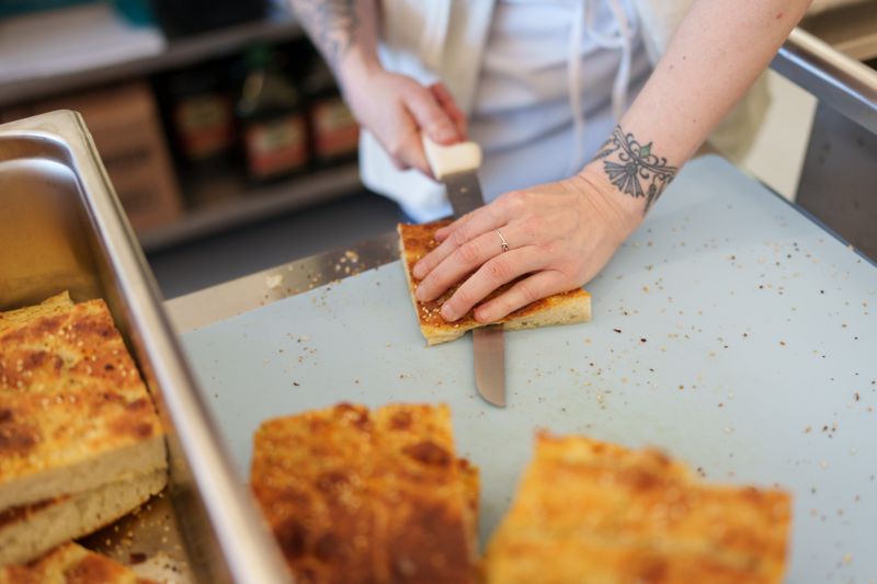 A hand with a tattoo around the wrist slicing a piece of focaccia bread on a cutting board, with other slices of bread visible in the foreground. 