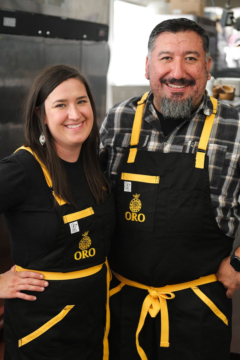 Chefs Kate and Gustavo Romero in black aprons with yellow ties facing the camera and smiling in the kitchen. 