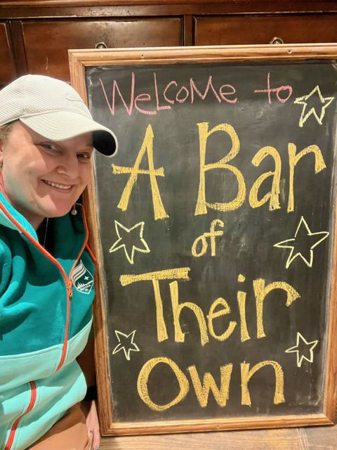 Jillian Hiscock, wearing a blue sweatshirt and white baseball cap, smiling next to a chalkboard with the words “Welcome to A Bar of Their Own” written on it in yellow chalk. 