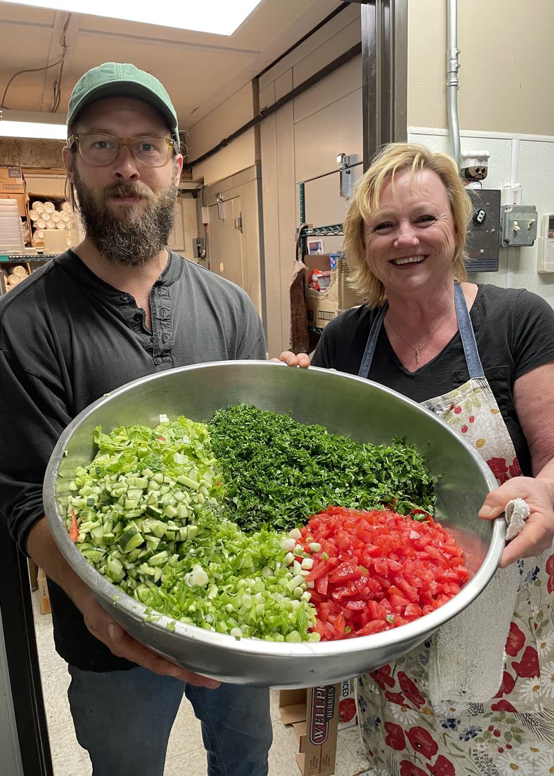 Samuel Miller and Ann Abukhodair standing in a kitchen, smiling and holding a large metal bowl full of chopped vegetables. 