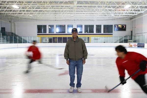 A  man stands on a hockey rink while two players skate around him