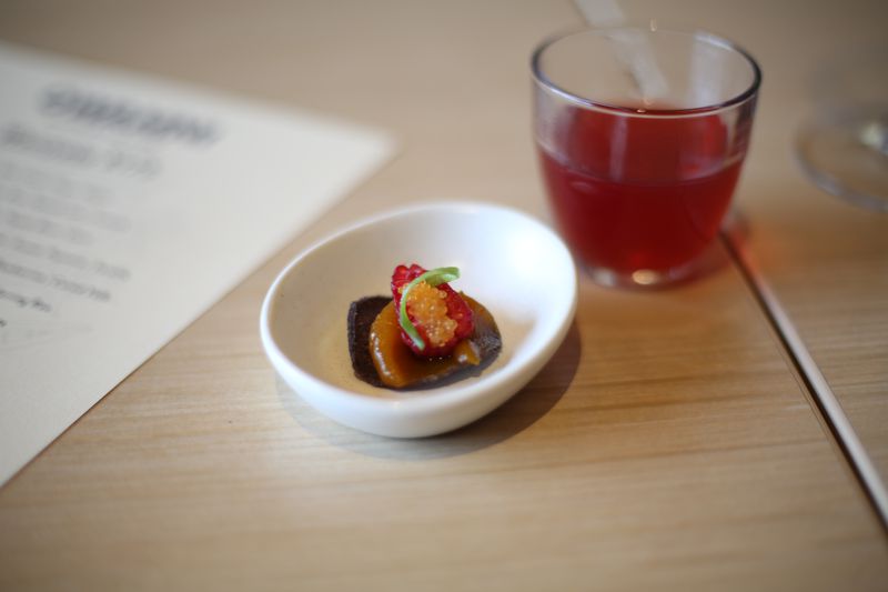 A small white clay dish with an oval blue corn chip topped with caramel-colored squash jam, a smoked raspberry, and golden trout roe. In the background, a small glass of bright red tea. To the left is a white printed menu with black lettering. 
