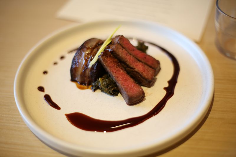 A white stone plate with three long and thing pieces of medium-rare bison meat beside a fatty ribeye cap over a bed of braised mushroom greens, with a drizzle of brown sauce in a circular shape on the plate, which is place on a light wooden table.