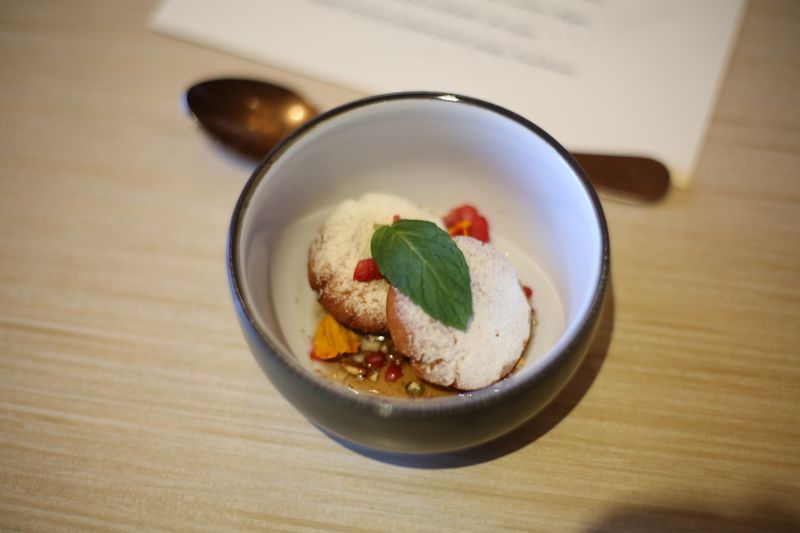 A small bowl with a brown exterior and white interior, holding two small doughnuts dusted with white powdered maple syrup, sitting in a light brown syrup, garnished with a mint leaf and small pieces of raspberries, all sitting on a light wooden table. 