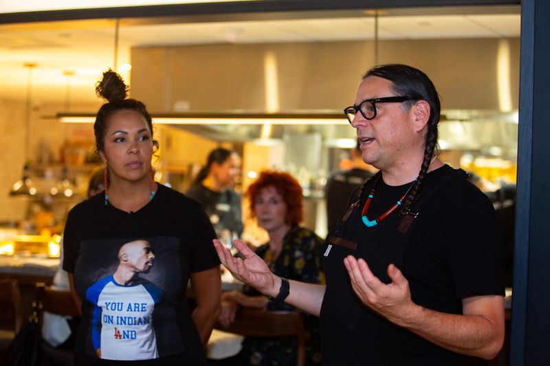 Mecca Boss, wearing a black T-shirt that says “You are on native land”, and Sean sherman, wearing a black T-shirt and a thick red and blue necklace, standing together and speaking to a crowd that’s off-camera. 