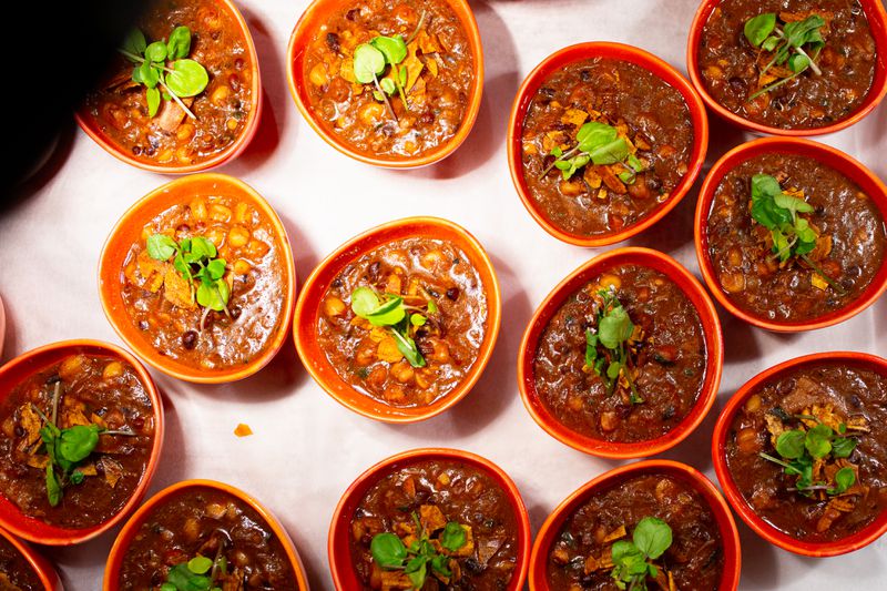 Orange bowls of a red, orange, and brown pumpkin stew, topped with sweet potato crisp and green herbs, on a white table, from an aerial point of view.