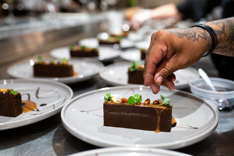 Rows of white plates with dark brown rectangular slices of cake, with a thin maple frosting and small delicate herbs on each; a tattooed hand reaching across the photo to place garnishes on top of a slice of the cake. 