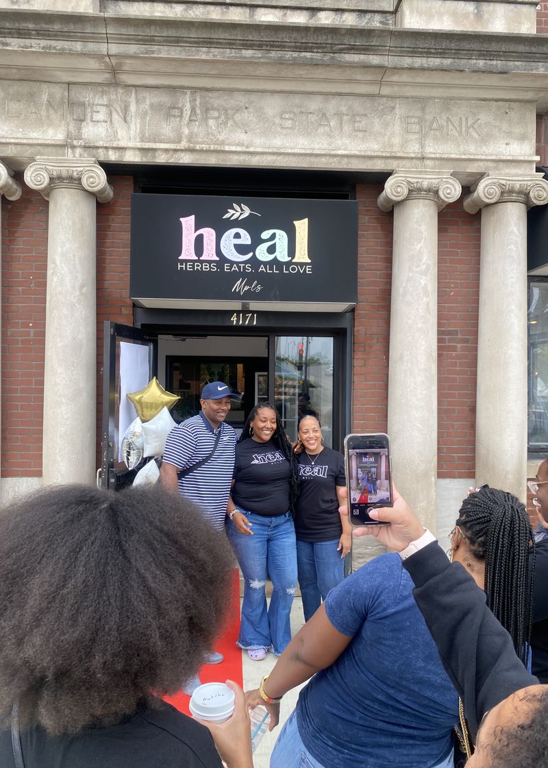 Sierra Carter wearing a black T-shirt and jeans standing with her barents in front of the Heal Mpls building, with concrete columns and a black sign that says “Heal” suspended above them. 