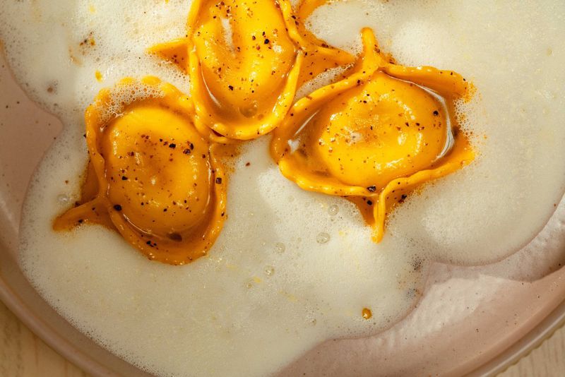 An up-close photo of three ravioli sitting on a light pink plate in a foamy sauce. 