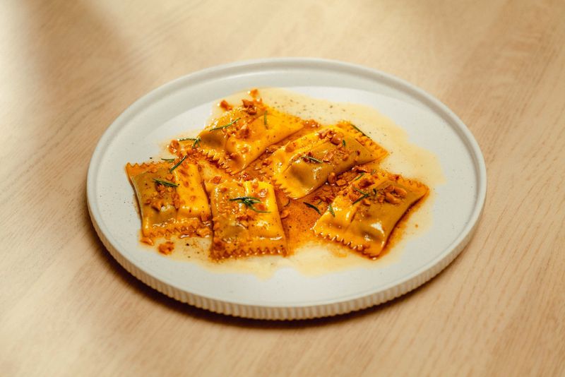 Five pieces of doppio ravioli, with two pockets and crimped edges, resting in a honey sauce on a round white plate on a blonde wood table. 