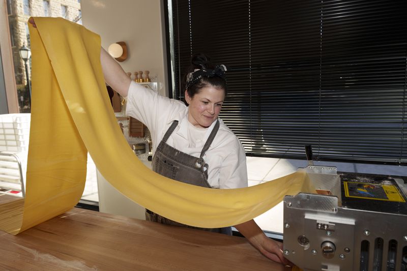 Rachel Cornelius McLeod, wearing a white shirt, gray apron, and black bandana in her hair, pulls a long pasta sheet out of a metal pasta machine, holding it up in the air.