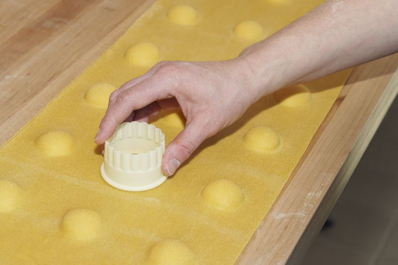 A hand pressing a cappelletti cutter with a scalloped edge onto a sheet of pasta with rounds of filling spaced evenly down its length.