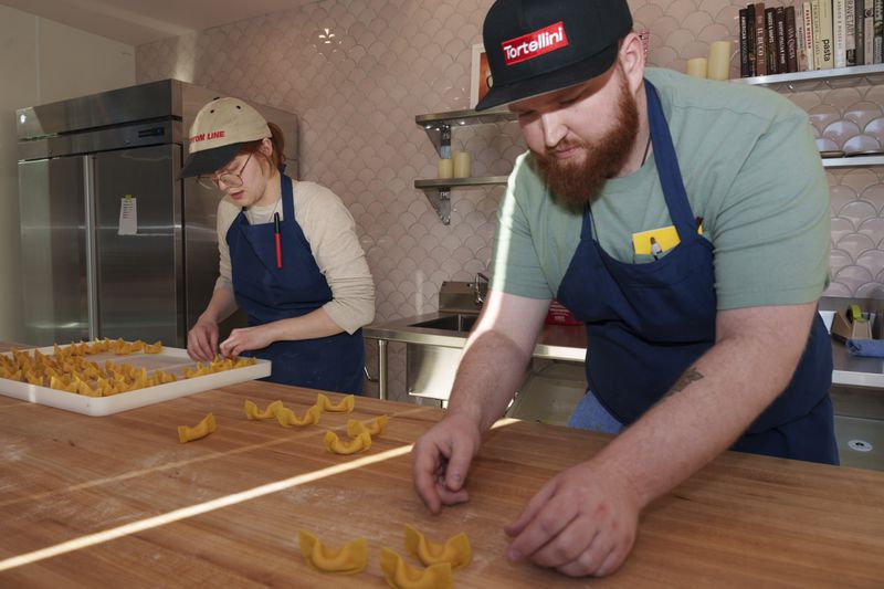 Two people wearing T-shirts, blue aprons, and baseball hats fold raviolis on a wooden table, with a pink tiled wall visible in the background. 