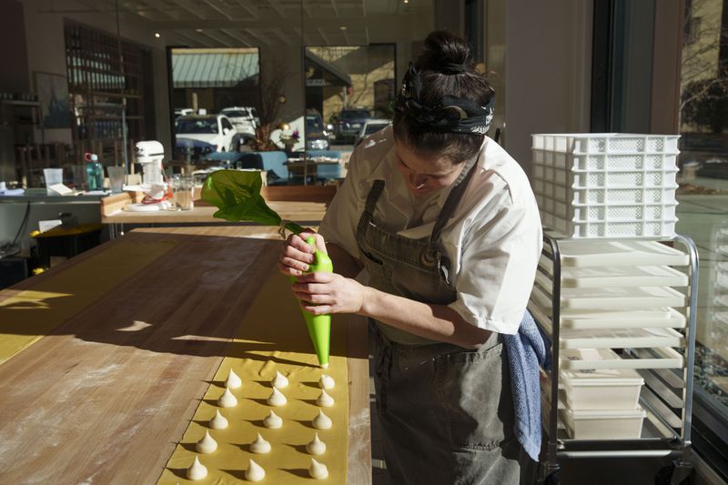 Rachel Cornelius McLeod piping dollops of white parmesan filling down a long pasta sheet on a narrow table, with pasta equipment and kitchen tools visible in the background. 