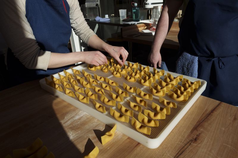A white mesh tray of scarpinocc pasta, and a person arranging a few scarpinocc on the corner of the sheet. 