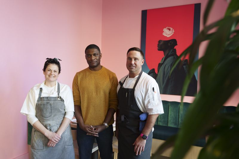 Rachel McLeod, Stephen Rowe, and Joe Rolle standing together in a pink room with a bright pink and dark blue painting on the wall, with plants visible in the foreground. 