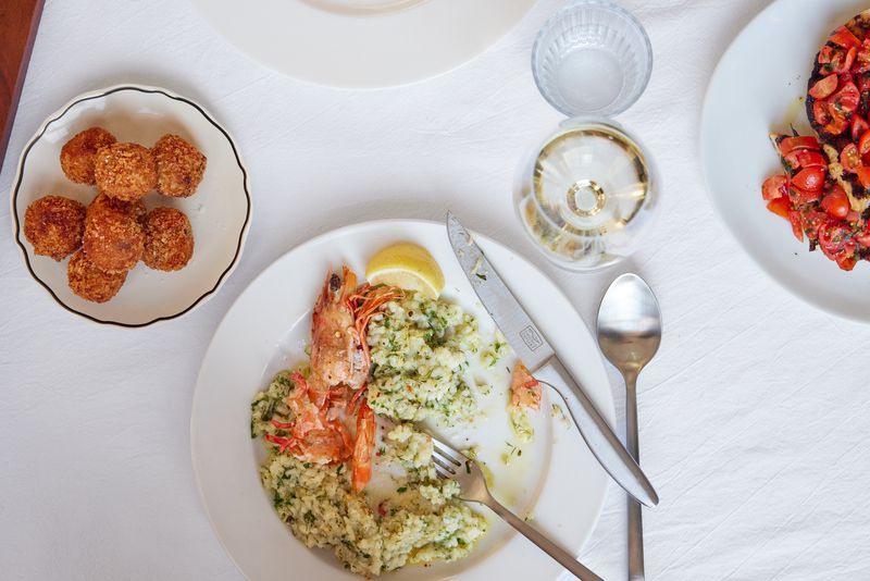 White plates of arancini, risotto with prawns, and tomato bruschetta on a white table cloth, with silverware and a wine glass. 