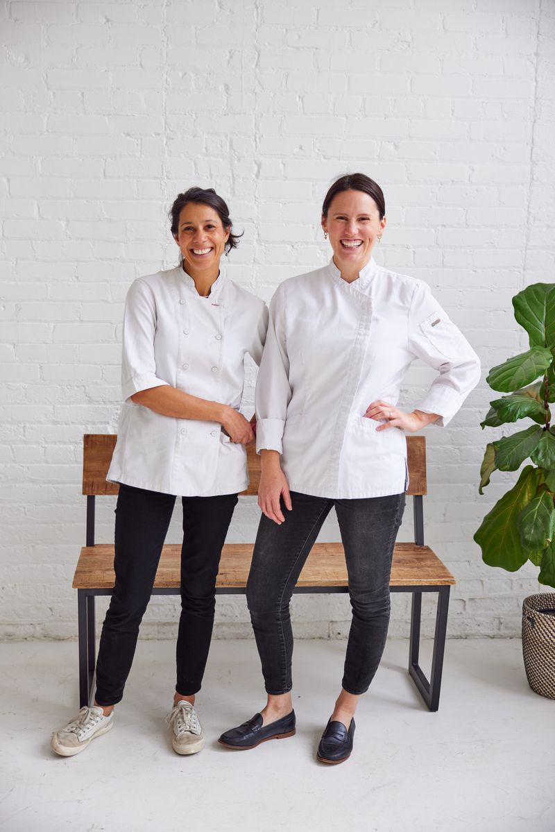 Chefs Jo Seddon and Lisa Wengler wearing white chefs coats and black pants, standing in front of a bench against a white wall, with a plant just to the right. 