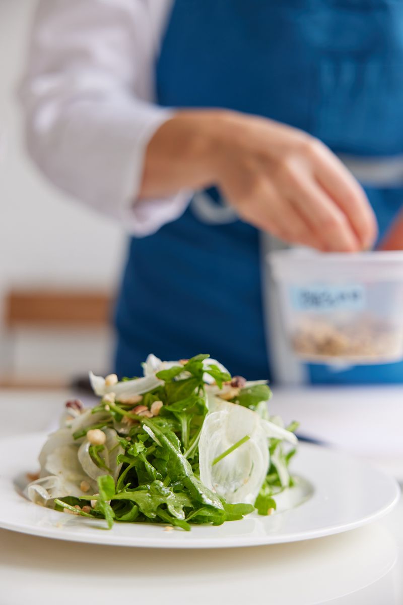 A white plate of arugula salad and a person in a white shirt and blue apron grabbing pine nuts out of a container behind it. 
