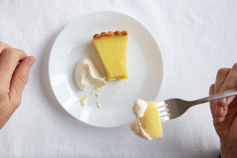 A slice of lemon tart on a white plate with a dollop of creme fraiche, two hands pulling a fork with a bite of the tart up toward the camera. 