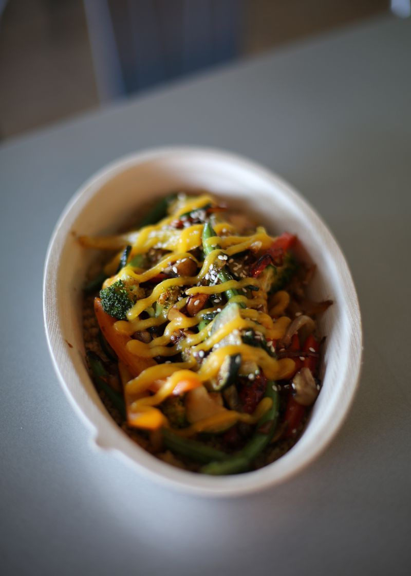 An oval bowl filled with vegetables topped with stripes of an orange sauce, sitting on a white table. 