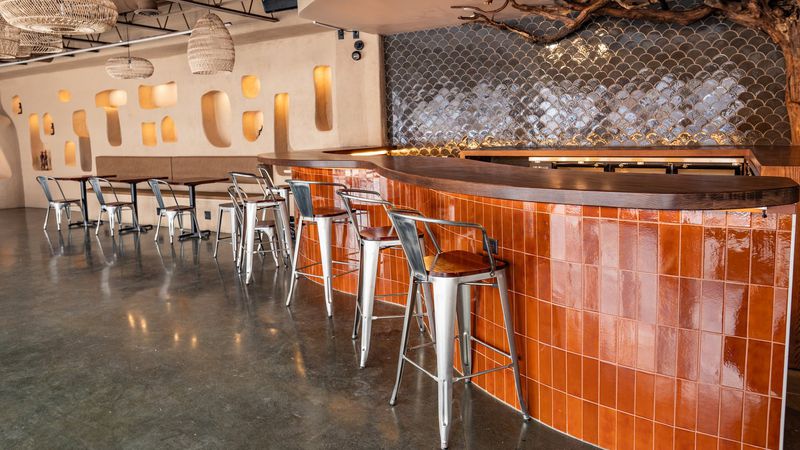 The interior of a restaurant with a curved bar with orange tiles underneath it, grey scalloped tiles and a driftwood sculpture behind the bar, and an earthy beige wall with sculptural alcoves. 