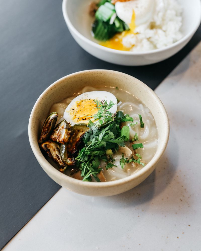 A beige ceramic bowl holding a noodle soup with chicken, egg, green herbs, and fried eggplant pieces. 