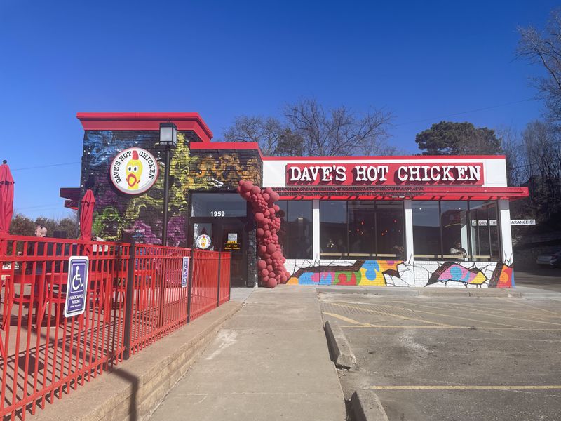 The exterior of Dave’s Hot Chicken, painted white and black with colorful murals and red accents, and a parking lot in the foreground. 