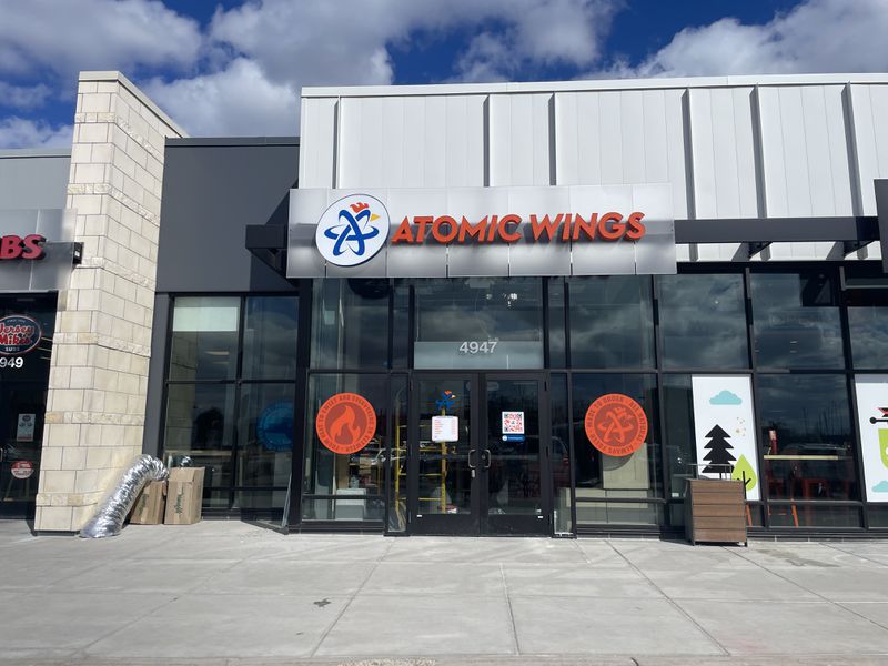 The exterior of a restaurant with large-paned glass windows and a sign that reads “Atomic Wings” in orange with sidewalk in the foreground and a blue sky with white clouds in the background. 