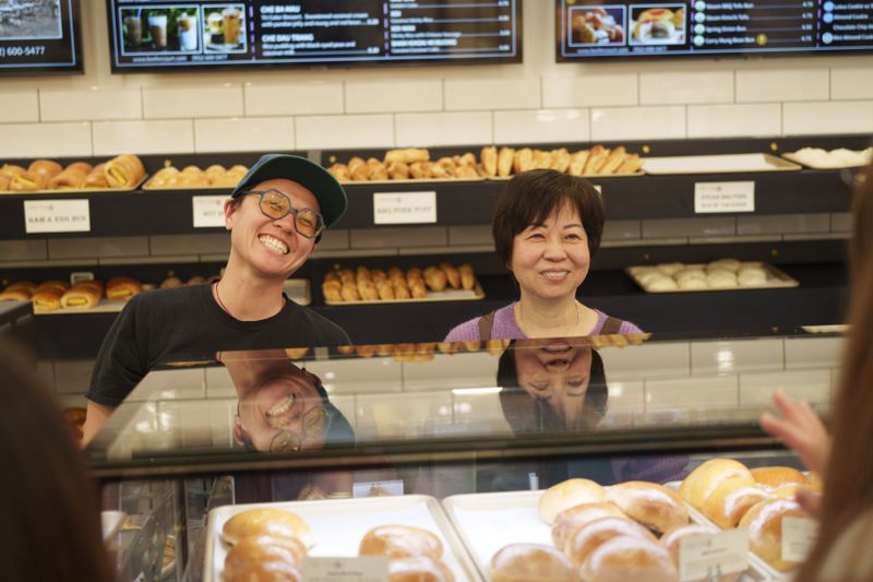 Michelle Kwan, wearing glasses and a baseball hat, and Paulina Kwan, wearing a purple sweater, stand behind the counter of Keefer Court Bakery and smile.