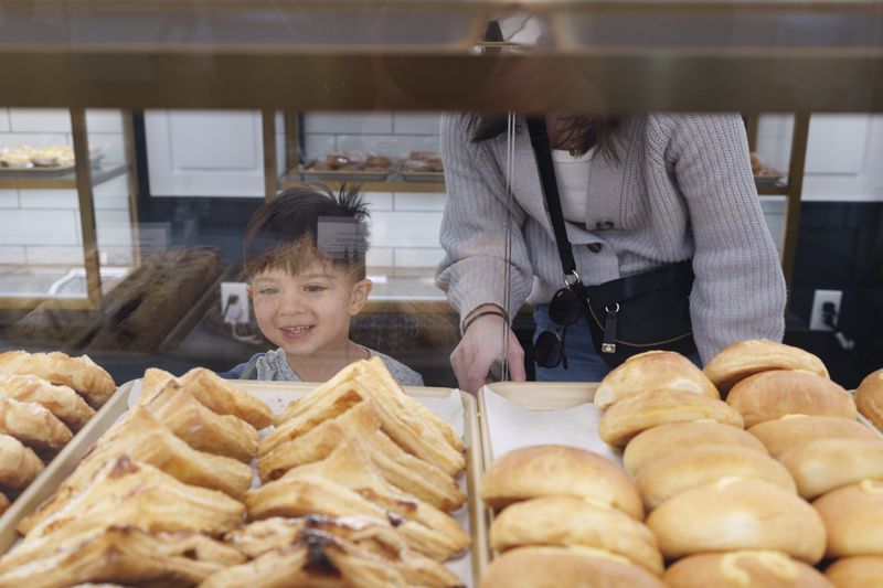 A small boy standing behind a glass pastry case and looking at the pastries in it a woman bending over him to his right.