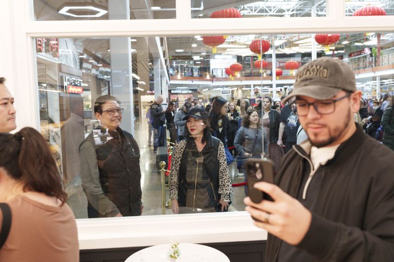 Behind the glass walls of the bakery, people stand in line as they wait to get in. In the foreground, a person with a beard, glasses, and a gray baseball hat takes a photo with his phone.