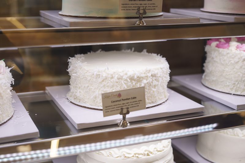A whole round frosted white cake with shredded coconut on the sides, sitting in a pastry case.