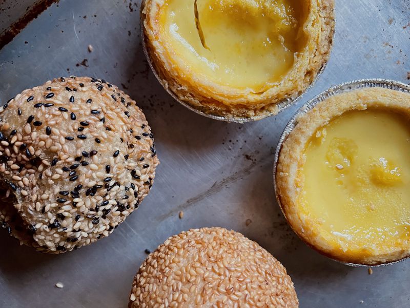 Two yellow egg tarts and two sesame balls on a metal surface. 