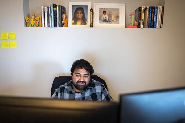 A man works at a desk, framed photos sit on a shelf above